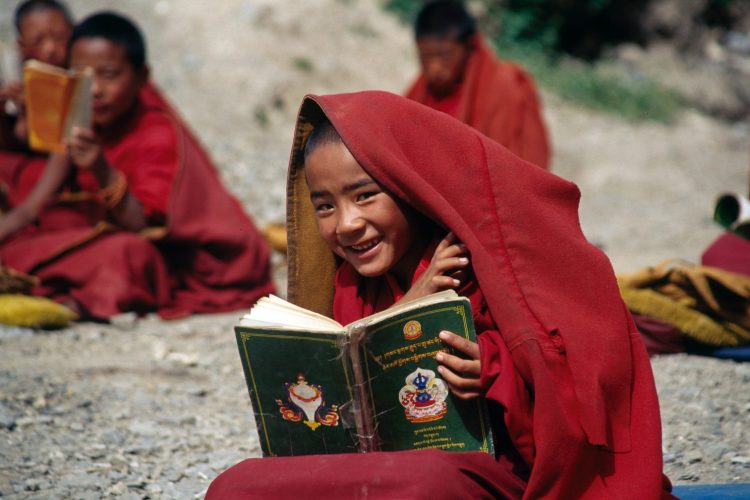 Seated Child Peeking from Red Hooded Robe with Open Book in Hand and Other Robed Children in Background